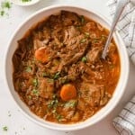 A white serving bowl full of homemade slow cooker beef stew, with a spoon in the bowl and chopped parsley on the side.